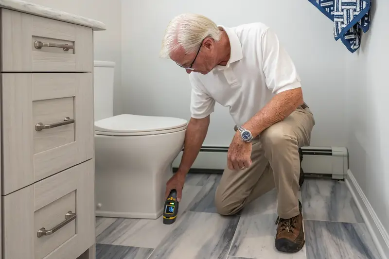 Service tech kneeling on a bathroom floor, checking around the base of a toilet with a handheld meter