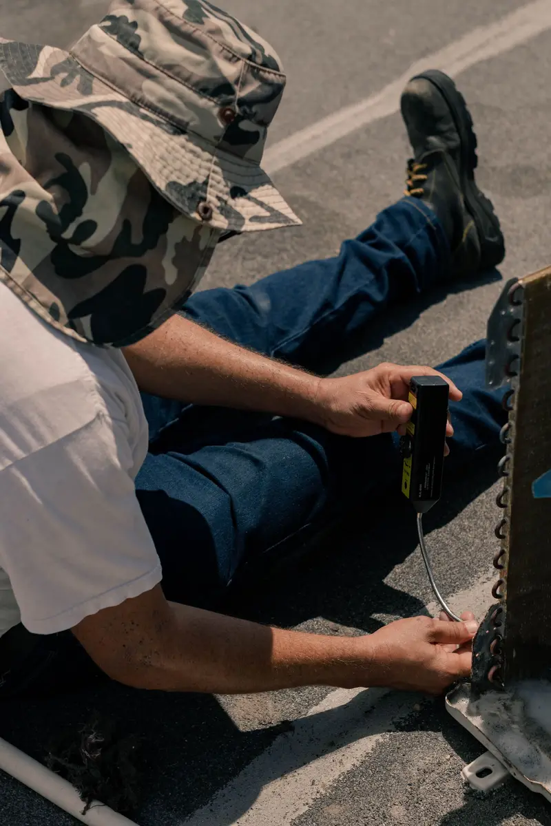AC technician sitting on a rooftop, running a refrigerant leak detector along a condenser coil